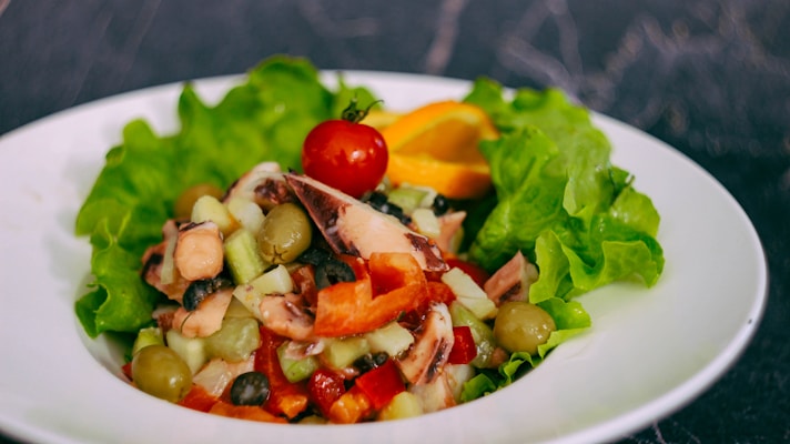 A colorful salad composed of fresh green lettuce, sliced vegetables, and a cherry tomato on top. There is an assortment of ingredients including olives, cucumbers, diced peppers, and possibly some form of seafood. The salad is presented on a white plate with a dark background.