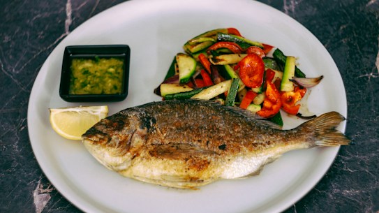 A grilled fish served on a white plate alongside a colorful assortment of roasted vegetables including zucchini, red bell peppers, and onions. A small square dish filled with green sauce and a lemon wedge are also present on the plate.