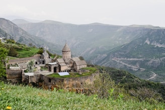 A historic stone monastery complex is perched on a lush green hillside, surrounded by expansive mountain ranges. The architecture features domed structures and arched doorways, typical of medieval design. The landscape is serene with greenery contrasting against the grey stone walls of the monastery. The background features rolling hills and steep mountain slopes shrouded in a hazy atmosphere.