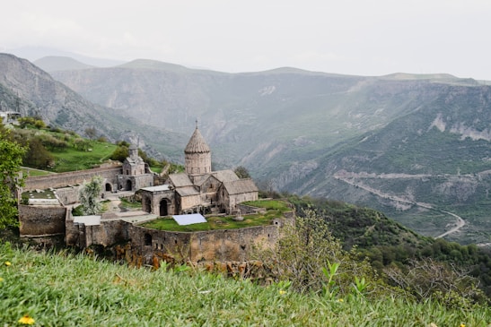 A historic stone monastery complex is perched on a lush green hillside, surrounded by expansive mountain ranges. The architecture features domed structures and arched doorways, typical of medieval design. The landscape is serene with greenery contrasting against the grey stone walls of the monastery. The background features rolling hills and steep mountain slopes shrouded in a hazy atmosphere.