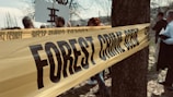 A yellow tape labeled 'FOREST CRIME SCENE' is wrapped around a tree, with several people gathered in the background and partially visible. The setting appears to be outdoors, with bare trees and a somewhat overcast sky.