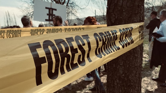 A yellow tape labeled 'FOREST CRIME SCENE' is wrapped around a tree, with several people gathered in the background and partially visible. The setting appears to be outdoors, with bare trees and a somewhat overcast sky.