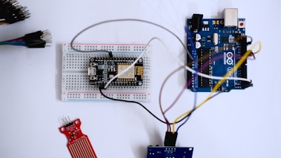 A microcontroller development board, breadboard, and various electronic components are arranged on a surface. Wires connect the components, indicating a circuit setup. The setting is clean with a focus on electronics and hardware prototyping.