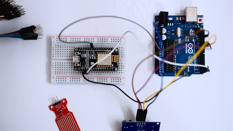 A microcontroller development board, breadboard, and various electronic components are arranged on a surface. Wires connect the components, indicating a circuit setup. The setting is clean with a focus on electronics and hardware prototyping.