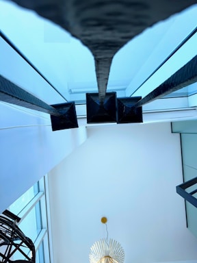 A modern interior space features large glass windows allowing natural light to flood in. Prominent black metal poles run vertically, adding a structural element. A gold, spiky chandelier hangs from the ceiling, creating an artistic focal point. The ceiling and walls are painted in white, enhancing the brightness of the room.
