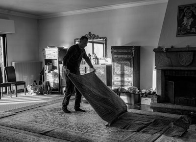 A person is rolling up a large patterned rug in a room filled with furniture and decorative items. The room includes a fireplace, chairs, a wooden dresser with intricate designs, a mirror, and a collection of small objects on and around the furniture. Natural light streams in from a window on the left, casting shadows across the floor.