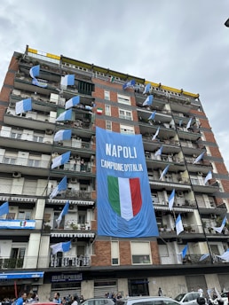 A multi-story residential building displays a large blue banner with 'NAPOLI CAMPIONE D'ITALIA' alongside an Italian flag emblem. Several balconies are adorned with blue and white flags, suggesting a celebratory atmosphere. The street below has people gathered, possibly participating in or observing a celebration.