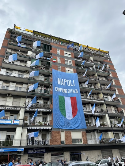 A multi-story residential building displays a large blue banner with 'NAPOLI CAMPIONE D'ITALIA' alongside an Italian flag emblem. Several balconies are adorned with blue and white flags, suggesting a celebratory atmosphere. The street below has people gathered, possibly participating in or observing a celebration.