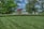 A well-manicured lawn extends across the foreground, bordered by a tidy row of hedges. Beyond the hedges, a variety of trees with lush green foliage and a few with red leaves create a natural screen. In the background, a body of water lies under a partly cloudy sky.