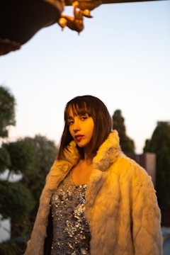 A person with shoulder-length brown hair is wearing a sequined dress and a light-colored fur jacket. The background features some greenery and a clear sky.