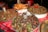A street food stall displays an array of exotic fried insects, snails, and seafood. Plates are piled high with crispy grasshoppers, sprinkled with green herbs and red chili peppers. In the background, there are large metal trays filled with cooked snails and fried shrimp.