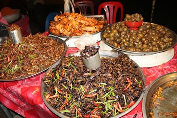 A street food stall displays an array of exotic fried insects, snails, and seafood. Plates are piled high with crispy grasshoppers, sprinkled with green herbs and red chili peppers. In the background, there are large metal trays filled with cooked snails and fried shrimp.