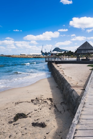 A wooden boardwalk runs alongside a sandy beach with some seaweed and waves gently hitting the shore. There is a stone retaining wall between the beach and the boardwalk. In the background, industrial buildings and cranes can be seen. A few structures with slanted roofs are visible near the boardwalk.