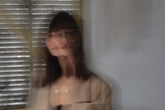 A blurred image of a woman standing in front of a window with horizontal blinds. The woman's face and upper body appear to be doubled or shifted, creating a disorienting effect. There is light coming through the blinds, and the background is visible through them.