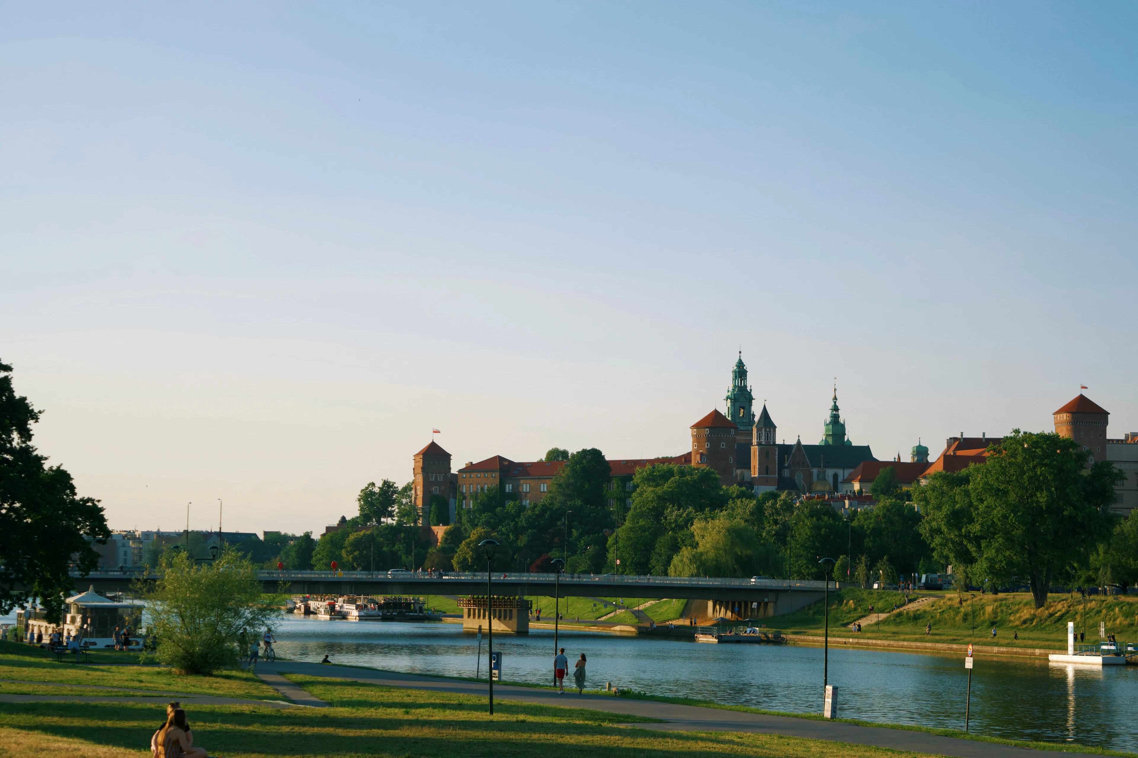Wawel Castle and its cathedral spires on the hill above the Vistula river in Krakow, seen across the water from the grassy riverside embankment.