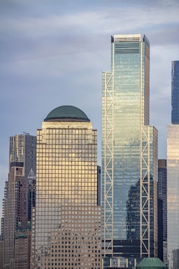 A cityscape featuring modern skyscrapers with reflective glass surfaces under a cloudy sky. The tallest building has a geometric, glass facade with diagonal lines, and another has a green dome on top.