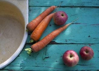 A rustic wooden table features three carrots and four red apples placed near a stone sink. The table has a weathered, turquoise paint, giving a vintage and homely feel. The carrots and apples are fresh, with vibrant colors contrasting against the weathered table.