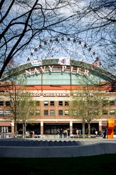 A large ferris wheel partially visible above brick buildings with the words 'NAVY PIER' prominently displayed. The building houses the Chicago Children's Museum. American flags and trees frame the view. People are casually walking and standing near the entrance.