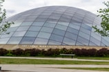 A large, dome-shaped building made of glass panels with metal framing, set in a landscaped area with green grass, bushes, and trees. A bench and a paved pathway are visible in the foreground. The building is identified as the Joe and Rika Mansueto Library.