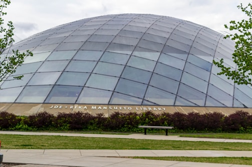 A large, dome-shaped building made of glass panels with metal framing, set in a landscaped area with green grass, bushes, and trees. A bench and a paved pathway are visible in the foreground. The building is identified as the Joe and Rika Mansueto Library.