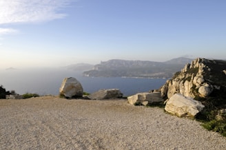A coastal landscape with a gravel path in the foreground, flanked by large white boulders. Beyond, the view extends to a vast body of water with distant islands barely visible on the horizon. Rugged cliffs and rolling hills frame the scene under a clear blue sky.