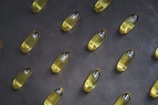 A collection of yellow capsules neatly arranged on a dark, textured surface. Each capsule is oblong and translucent, containing an oily liquid.