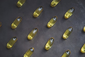 A collection of yellow capsules neatly arranged on a dark, textured surface. Each capsule is oblong and translucent, containing an oily liquid.