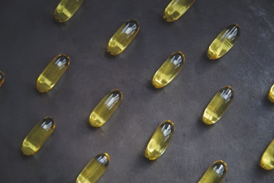A collection of yellow capsules neatly arranged on a dark, textured surface. Each capsule is oblong and translucent, containing an oily liquid.