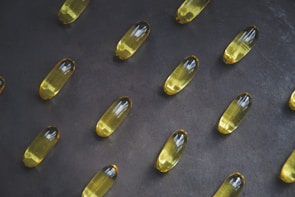 A collection of yellow capsules neatly arranged on a dark, textured surface. Each capsule is oblong and translucent, containing an oily liquid.