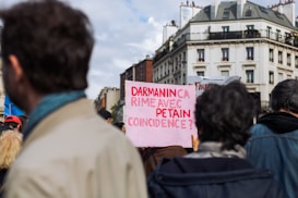 A group of people gathered in a city setting, holding various protest signs. One prominent sign reads, 'DARMANIN CA RIME AVEC PETAIN COINCIDENCE?' in red letters on a pink background. The crowd is viewed from behind, and buildings are visible in the background under a partly cloudy sky.