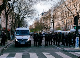 A city street lined with tall trees and historical buildings is occupied by police officers in riot gear standing in front of white police vans. The atmosphere suggests a controlled public order situation with officers wearing helmets and shields, indicating preparedness for potential crowd management. The street is relatively empty otherwise, with visible debris on the ground and traffic lights showing a green pedestrian signal.