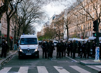 A city street lined with tall trees and historical buildings is occupied by police officers in riot gear standing in front of white police vans. The atmosphere suggests a controlled public order situation with officers wearing helmets and shields, indicating preparedness for potential crowd management. The street is relatively empty otherwise, with visible debris on the ground and traffic lights showing a green pedestrian signal.