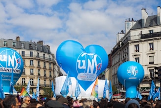 A street scene featuring large blue balloons with logos affiliated with a union or organization, surrounded by a crowd of people. The backdrop consists of classic European architecture with multi-story buildings and a partly cloudy sky. Colorful flags are visible, enhancing the sense of a public gathering or event.