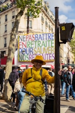 A person is standing outdoors holding a colorful protest sign that reads 'NE PAS BATTRE EN RETRAITE POUR SAUVER NOS RETRAITES'. The individual is wearing a yellow jacket, hat, and glasses, and is beside a bicycle. In the background, there are other people walking and buildings with shops and trees lining the street.