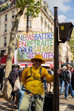 A person is standing outdoors holding a colorful protest sign that reads 'NE PAS BATTRE EN RETRAITE POUR SAUVER NOS RETRAITES'. The individual is wearing a yellow jacket, hat, and glasses, and is beside a bicycle. In the background, there are other people walking and buildings with shops and trees lining the street.