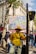 A person is standing outdoors holding a colorful protest sign that reads 'NE PAS BATTRE EN RETRAITE POUR SAUVER NOS RETRAITES'. The individual is wearing a yellow jacket, hat, and glasses, and is beside a bicycle. In the background, there are other people walking and buildings with shops and trees lining the street.