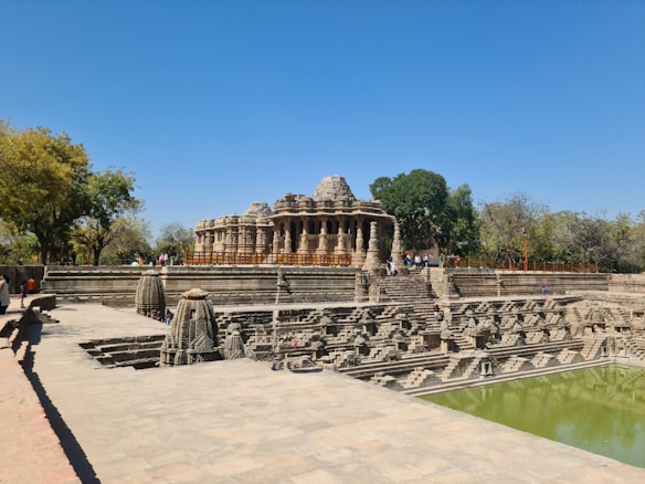 An ancient temple with intricate stone carvings stands against a bright blue sky. The structure is surrounded by steps leading down to a water tank, which reflects the temple. Trees and scattered visitors add to the scene.