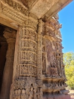 Intricate stone carvings adorn a historical temple facade, featuring numerous detailed sculptures and patterns depicting human figures and mythical creatures. The carvings, highlighted by natural sunlight, exhibit remarkable craftsmanship with a blend of ornate geometric and floral designs.