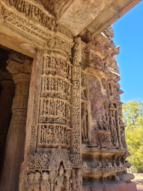 Intricate stone carvings adorn a historical temple facade, featuring numerous detailed sculptures and patterns depicting human figures and mythical creatures. The carvings, highlighted by natural sunlight, exhibit remarkable craftsmanship with a blend of ornate geometric and floral designs.