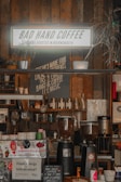 Interior of a cozy coffee shop with rustic wooden walls. A sign reads 'Bad Hand Coffee,' surrounded by various plants and coffee-related equipment. Several coffee grinders and an espresso machine are visible on the counter, along with a variety of small jars and containers. The atmosphere appears casual and inviting, ideal for enjoying a cup of coffee.