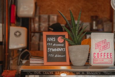 A small chalkboard sign offers a 15% discount for NHS workers and students. The sign is placed on a counter beside a potted plant with long, pointed green leaves. A bag of 'South Coast Coffee' is to the right of the plant. The scene is set in a cozy coffee shop environment with stacks of items in the background.