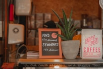 A small chalkboard sign offers a 15% discount for NHS workers and students. The sign is placed on a counter beside a potted plant with long, pointed green leaves. A bag of 'South Coast Coffee' is to the right of the plant. The scene is set in a cozy coffee shop environment with stacks of items in the background.