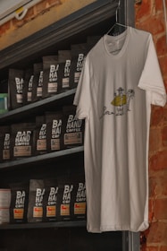 A white t-shirt with a printed cartoon design hangs on a wall. The design features a person holding a drink, standing next to a palm tree and a small creature. Beneath the shirt is a shelf stocked with bags of coffee, each labeled 'Bad Hand Coffee' with different roast names like French Roast and Morning Blend. The setting has a rustic feel with a brick wall partially visible.