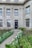 An elegant building façade featuring a dark door framed by classical architectural details, with tall windows above and on either side. A path lined with blooming white flowers and green shrubs leads to the entrance, creating a welcoming atmosphere.