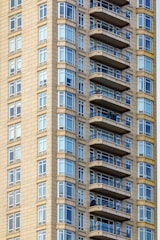 A close-up view of a modern high-rise building featuring numerous balconies and large glass windows. The exterior is tiled with a beige material, providing a sleek, contemporary appearance.