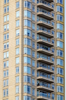 A close-up view of a modern high-rise building featuring numerous balconies and large glass windows. The exterior is tiled with a beige material, providing a sleek, contemporary appearance.