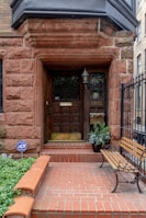 An entrance to a building featuring a wooden door with decorative glass panels and a brass mail slot. Surrounding the door is a stone frame made of reddish-brown bricks, with a black lantern affixed to the wall. A potted plant is placed beside the door, and a wooden bench with iron armrests is set on the right side. The entryway is bordered by a low brick wall and a black wrought iron fence. A blue security sign is visible among the foliage.
