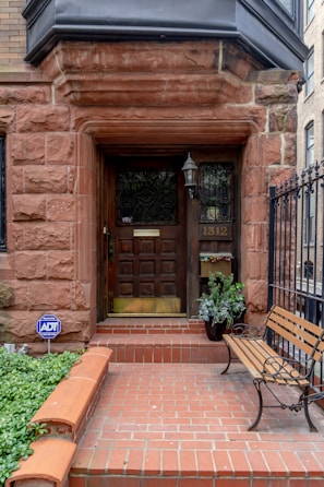 An entrance to a building featuring a wooden door with decorative glass panels and a brass mail slot. Surrounding the door is a stone frame made of reddish-brown bricks, with a black lantern affixed to the wall. A potted plant is placed beside the door, and a wooden bench with iron armrests is set on the right side. The entryway is bordered by a low brick wall and a black wrought iron fence. A blue security sign is visible among the foliage.
