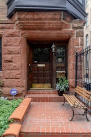 An entrance to a building featuring a wooden door with decorative glass panels and a brass mail slot. Surrounding the door is a stone frame made of reddish-brown bricks, with a black lantern affixed to the wall. A potted plant is placed beside the door, and a wooden bench with iron armrests is set on the right side. The entryway is bordered by a low brick wall and a black wrought iron fence. A blue security sign is visible among the foliage.