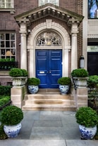 A grand entrance featuring a striking blue double door set within an ornate stone archway. The entrance is accentuated by neatly trimmed topiary plants placed in decorative blue and white porcelain pots. The brick facade of the building provides a classic and elegant backdrop, and a small set of stone stairs leads up to the door.